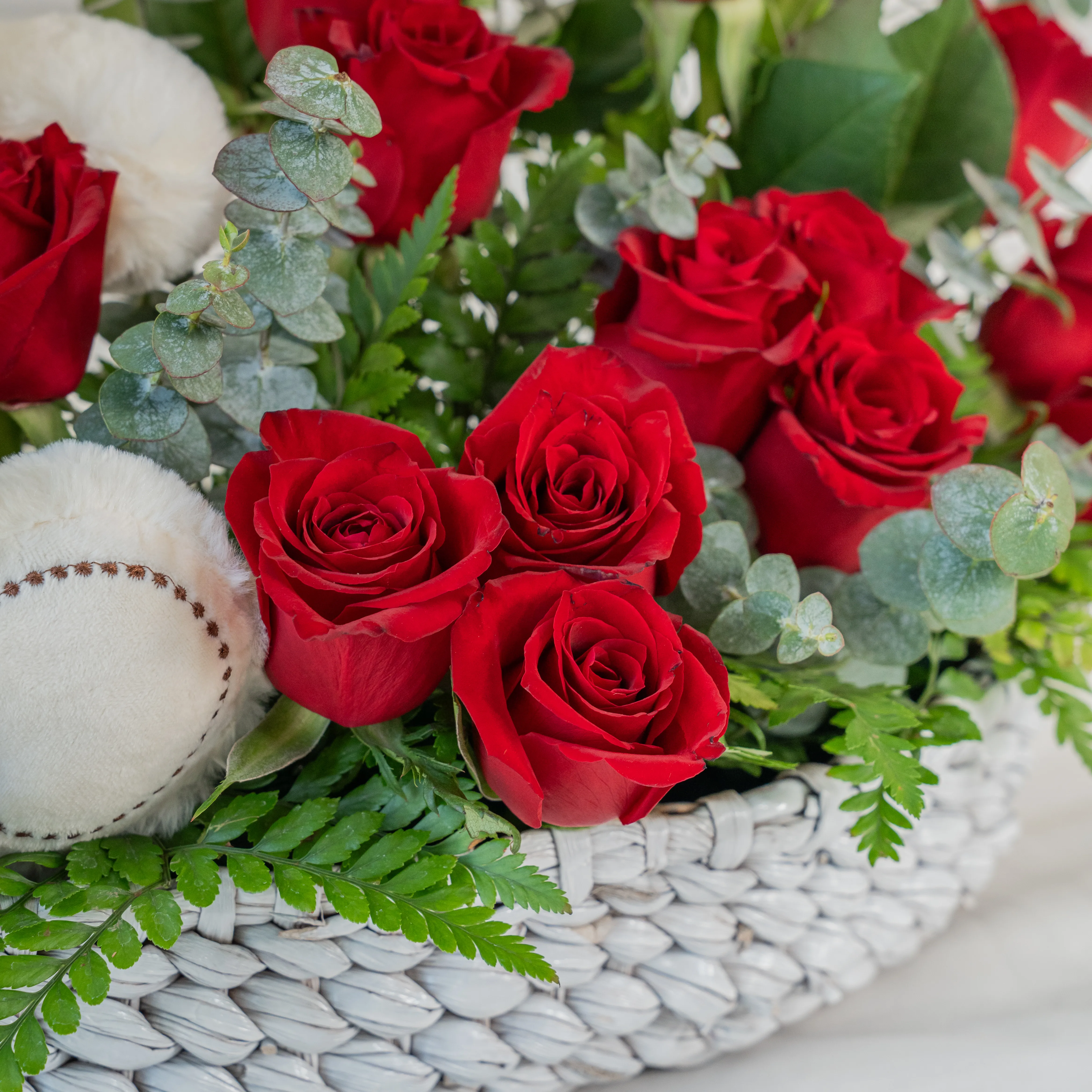 Basket arrangement of 20 red roses and small white teddy bear with eucalyptus leaf and green fillers.