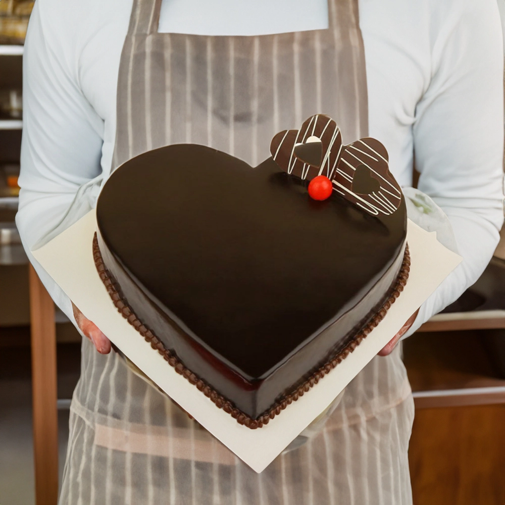 Heart shaped 1 kg chocolate truffle cake with cherry and chocolate hearts held by a person in an apron