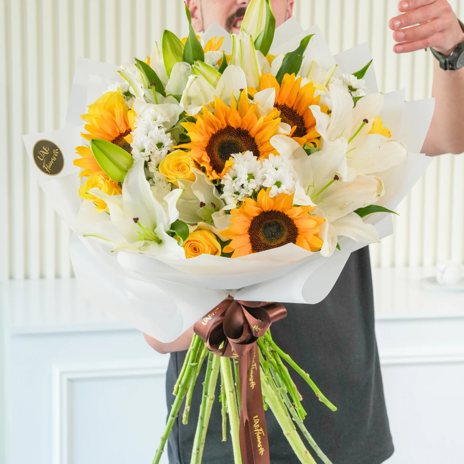 Bright bouquet of sunflowers, white lilies, yellow roses, and white filler flowers wrapped in elegant white paper with a brown satin ribbon – joyful floral gift by UAE Flowers.