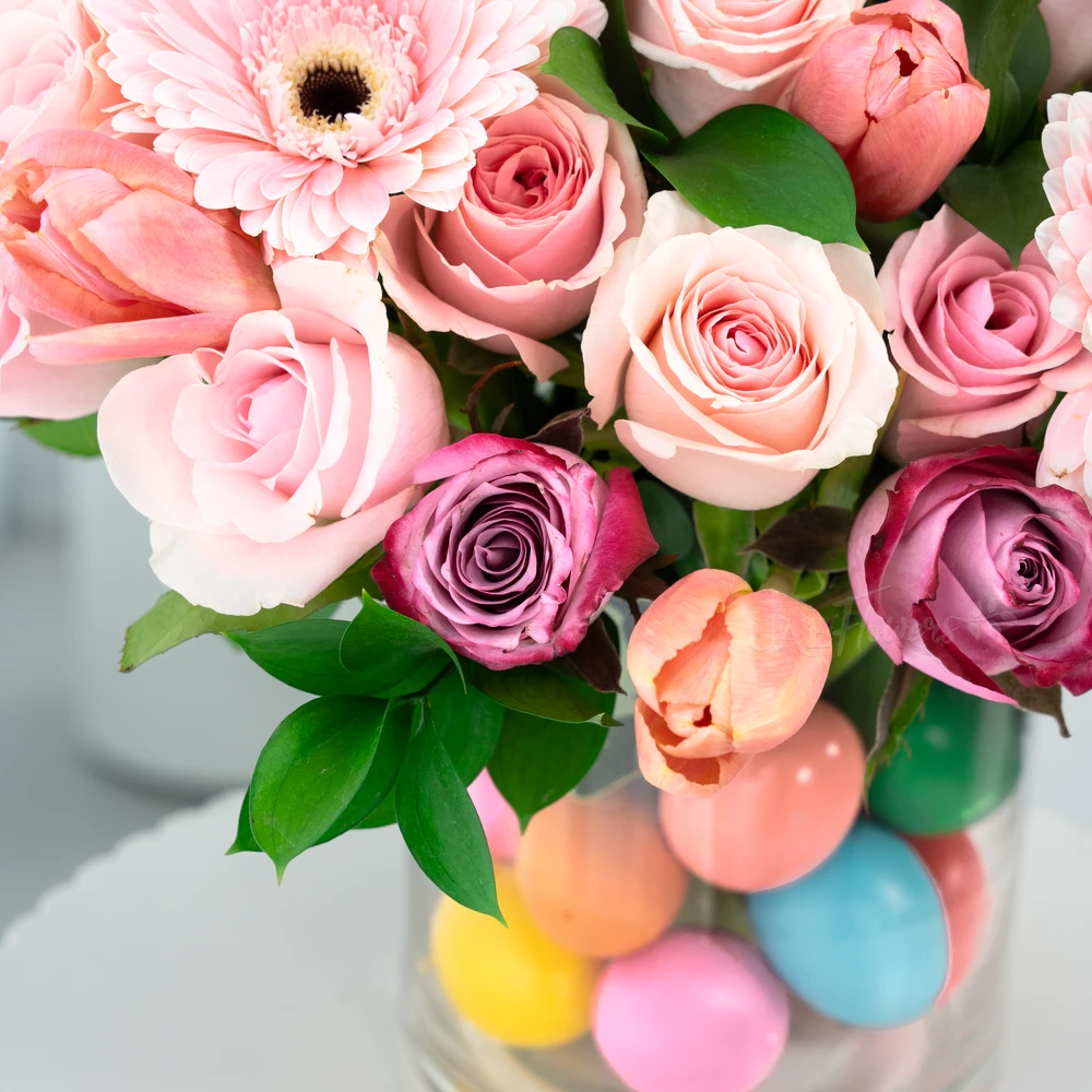 A vase arrangement of 3 pink gerberas, 7 purple roses, 15 pink roses, 10 pink tulips with green fillers and multi colored easter eggs.