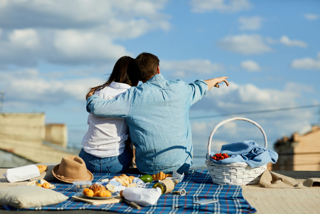 Young man embracing his girlfriend while sitting on roof and pointing at city scape